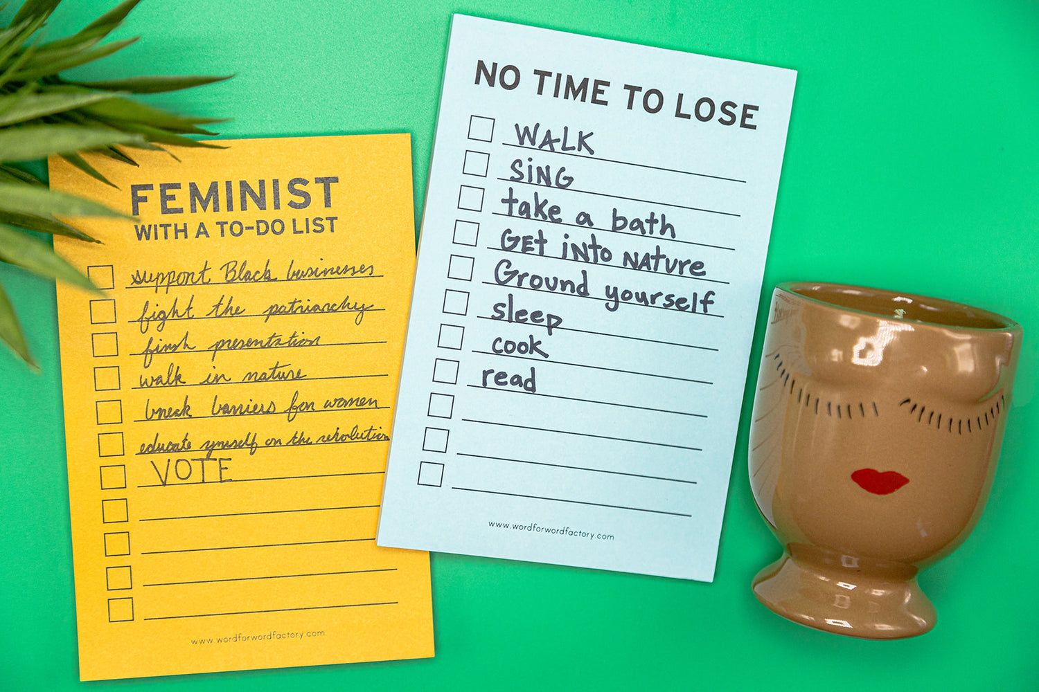 A gold notepad that said "Feminist with a to do list" a blue notepad that says "no time ot lose" and a clay mug with eyelashes and red lips